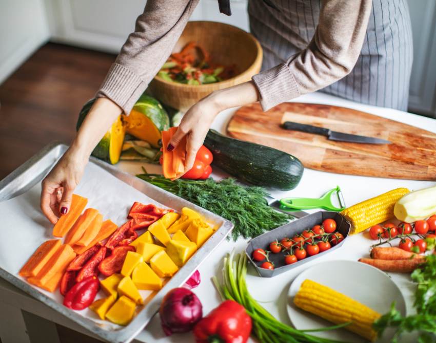 Oberkörper und Hände die Karotten auf einem Tablett anrichten, auf dem bereits gelbe und rote Paprika liegen. Auf dem Tisch liegen noch Zuchchini und Tomaten und ein Messer und Schneidbrett.