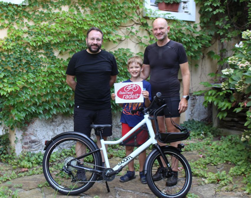 2 Männer, 1 Kind stehen hinter einem Kinderfahrrad, das Kind hat das Österreich radelt Logo in A4-Größe in der Hand