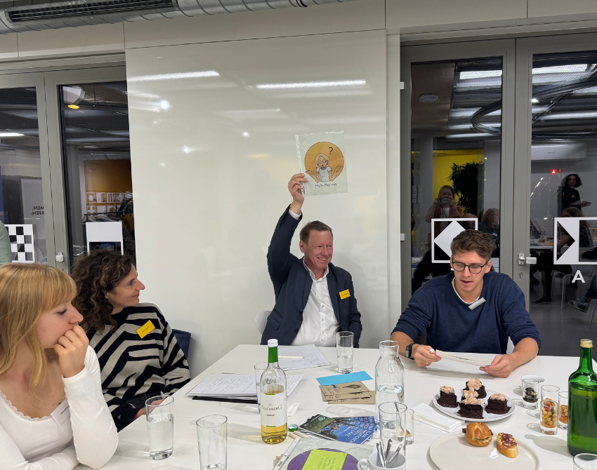 Gruppe sitzt am Tisch und Mann hält A4 Bild hoch mit Grafik einer Frau