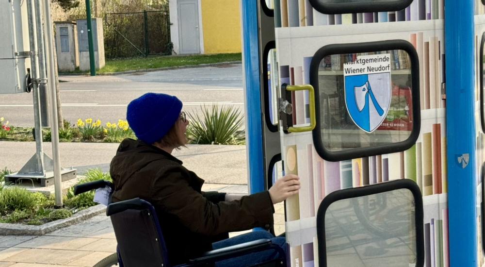 Person im Rollstuhl öffnet eine öffentliche Bücherzelle